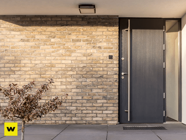 Moderne Haustür aus dunklem Material in Ziegelsteinfassade mit kleiner Pflanze davor.